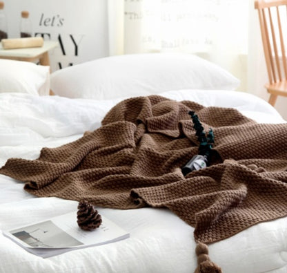 Brown knitted blanket on a white bed with a book and decorative items.