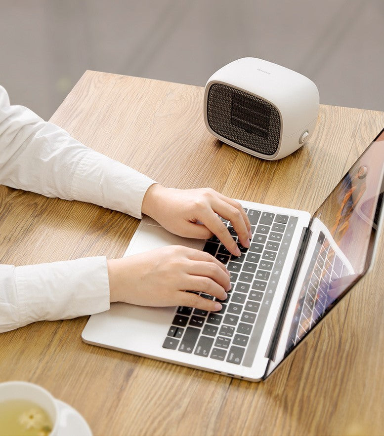 Person using a laptop with a small white heater on a wooden desk