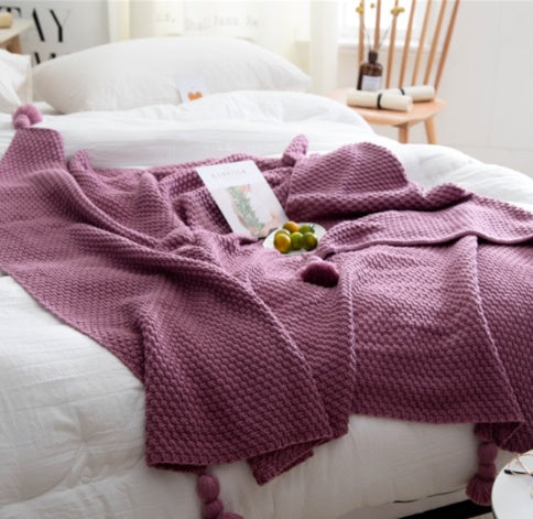 Purple knitted blanket on a bed with a white pillow and small plant in a room.