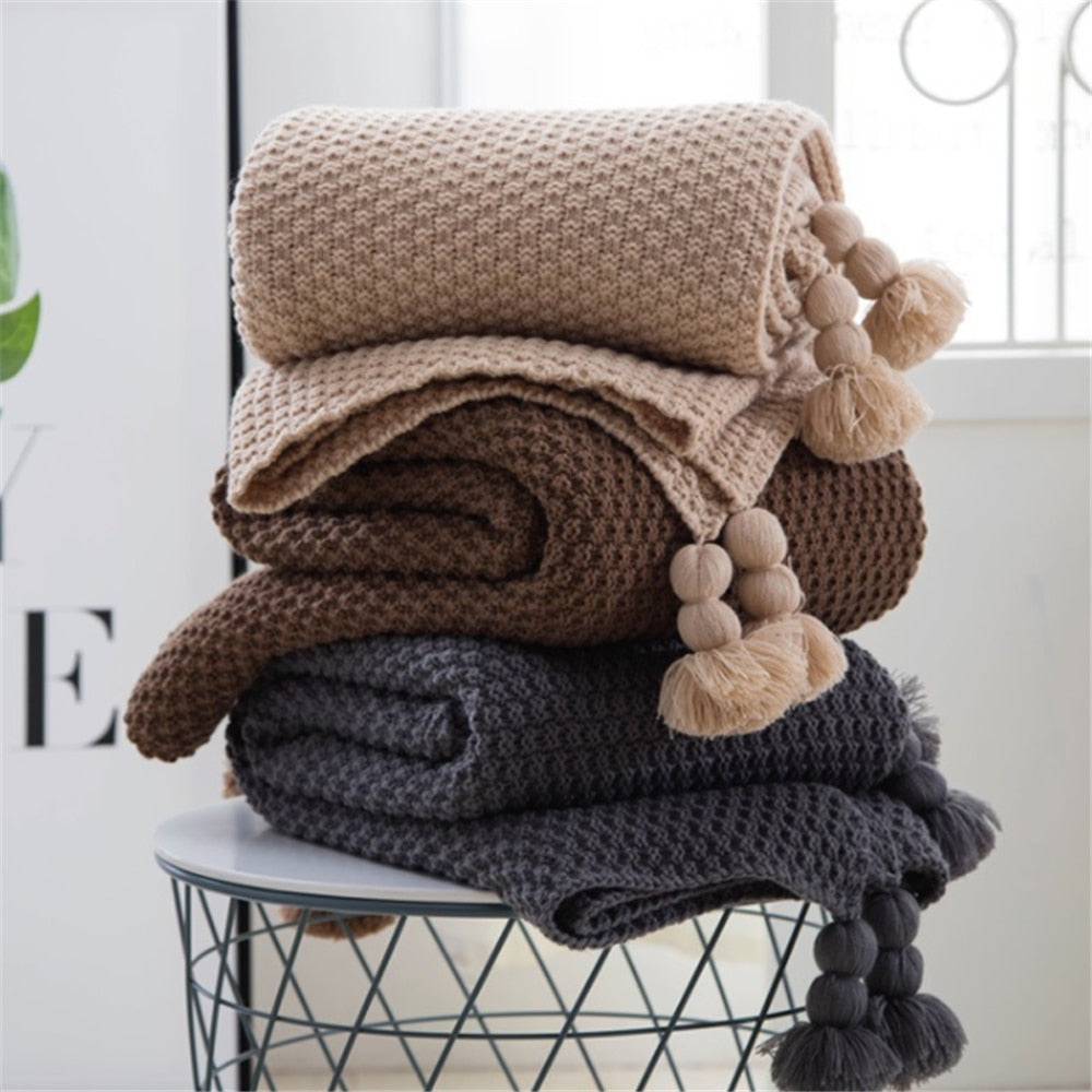 Stack of knitted blankets with pom-poms on a chair against a white wall.